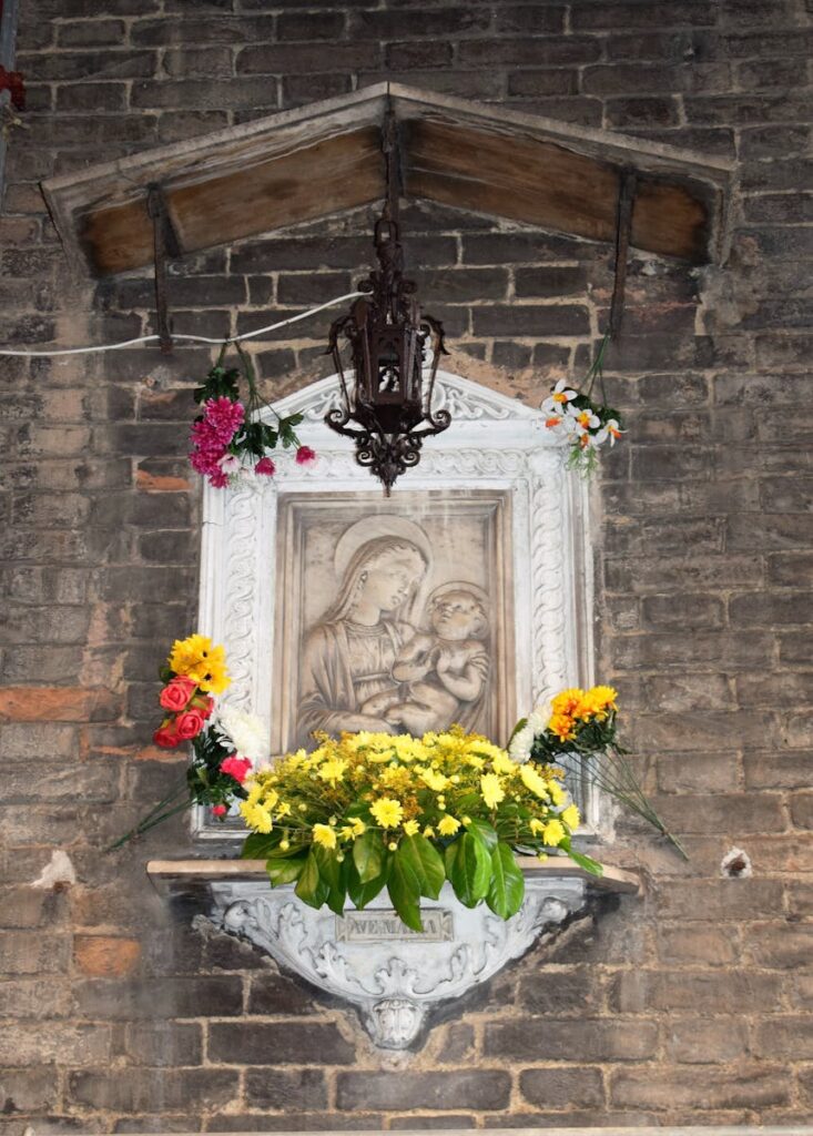 A decorative altar of Virgin Mary and Child adorned with fresh flowers on a brick wall in Siena.