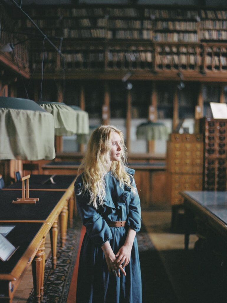 Elegant blonde woman in a blue dress standing in a vintage library with classic lighting.